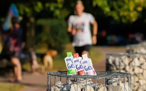 Trzy opakowania żeli energetycznych dla biegaczy: zielone jabłko, cola i truskawka z bananem, na tle biegnącej osoby.