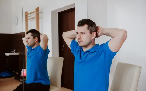 Mężczyzna w niebieskiej koszulce wykonuje ćwiczenia na szyję, opierając dłonie o tył głowy.