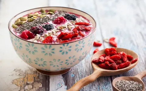 Miseczka smoothie z owocami, nasionami chia, pestkami dyni i słonecznika. Obok łyżki z jagodami goji i nasionami chia. Pyszny sposób, jak jeść nasiona chia.