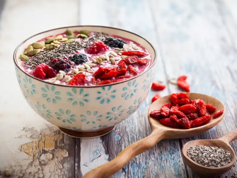 Miseczka smoothie z owocami, nasionami chia, pestkami dyni i słonecznika. Obok łyżki z jagodami goji i nasionami chia. Pyszny sposób, jak jeść nasiona chia.