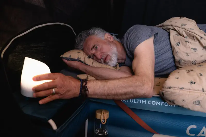 Mężczyzna z bólem próbuje znaleźć pozycję do spania przy rwie kulszowej, opierając się na poduszce i trzymając lampkę.
